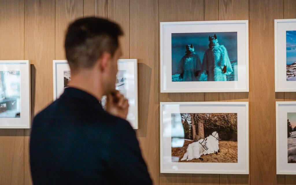 Eine Person in einem Anzug betrachtet nachdenklich gerahmte Fotos an einer Holzwand, die surreale Bilder von Figuren in pelzigen Kostümen und einem Pferd zeigen. Die Kulisse wirkt kontemplativ.