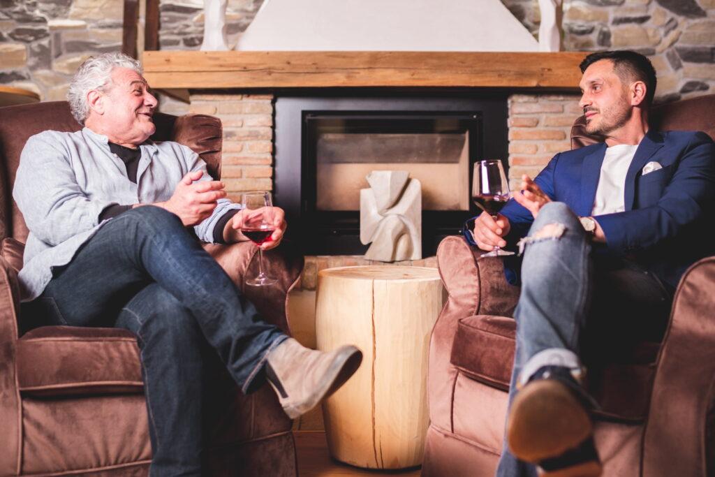 Zwei Männer sitzen gemütlich in Sesseln am Kamin im veganen Kunsthotel Paradiso Pure.Living in Südtirol und halten Weingläser in den Händen. Sie unterhalten sich angeregt und schaffen so eine warme, entspannte Atmosphäre.