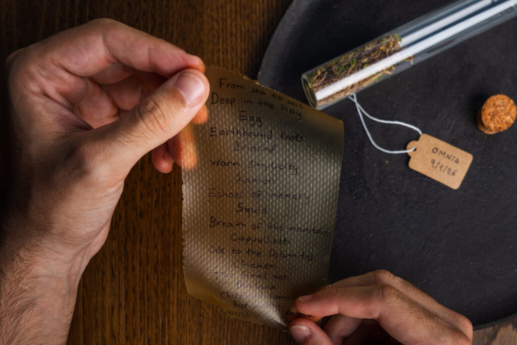 Zwei Hände halten ein strukturiertes Papier mit handgeschriebenen Worten. Ein Glasröhrchen mit Kräutern steht auf einer schwarzen Oberfläche neben einem Korken und einem beschrifteten Etikett und sorgt für eine rustikale Atmosphäre. Menü aus dem veganen Fine-Dining Restaurant OMNIA in Südtirol