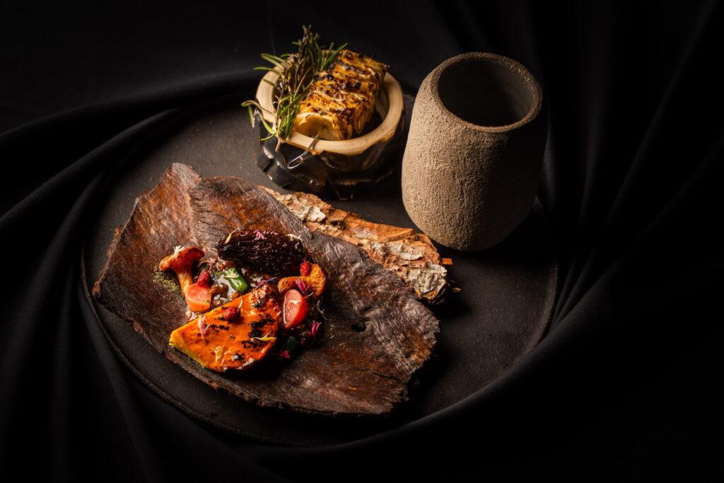 Exquisites veganes Gericht auf einem rustikalen Holzteller mit knackigem Gemüse, daneben gegrilltes Brot in einer steinartigen Schale und eine strukturierte Tasse auf einem schwarzen Tuch. Aus dem veganen Fine Dining Restaurant OMNIA