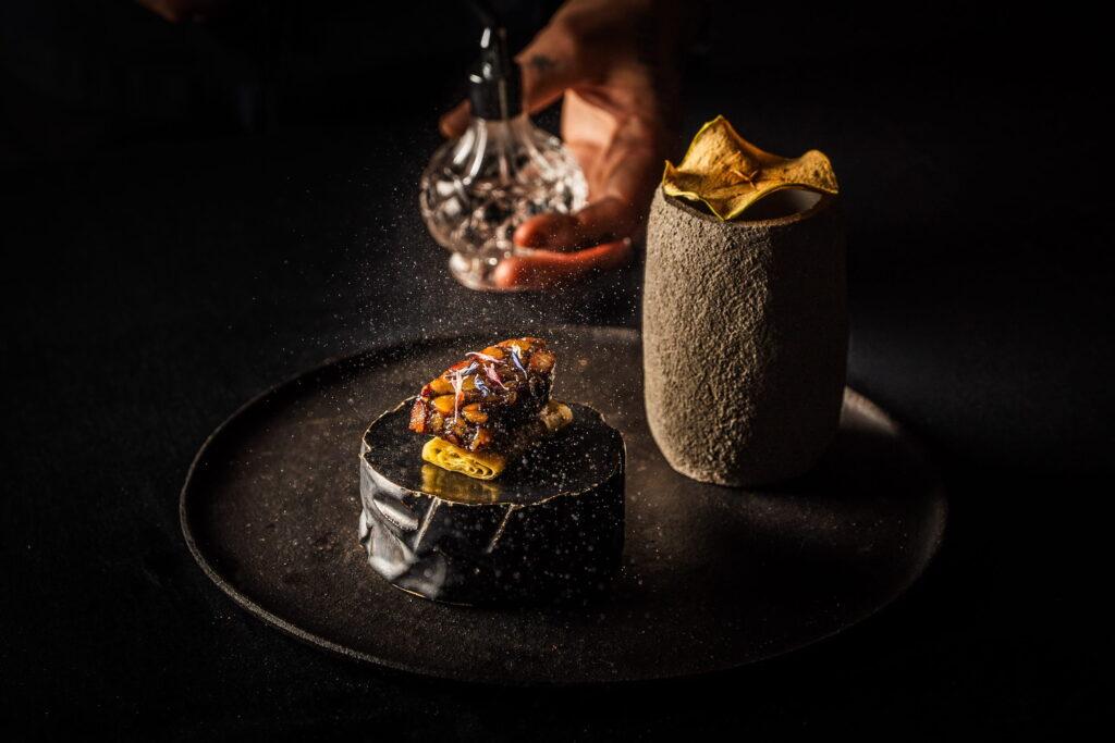 Elegantes veganes Gericht auf einem dunklen Teller, das eine Gourmet-Delikatesse und eine strukturierte Tasse präsentiert. Eine Hand streut Gewürze darüber und schafft so eine raffinierte, künstlerische Atmosphäre. Entstanden im OMNIA plant-based restaurant