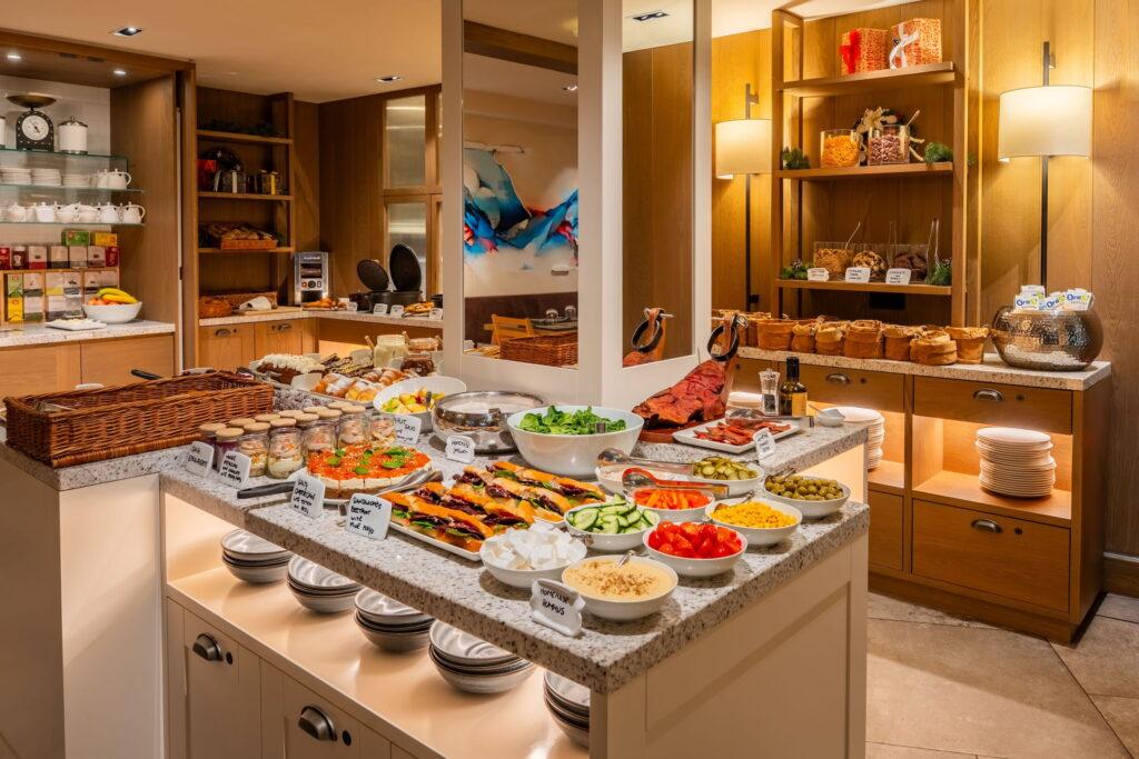 Veganes Buffet im veganen Südtiroler Hotel Paradiso Pure.Living mit bunten Salaten, Aufschnitt, Käse und Brot in einem warm beleuchteten Raum. Regale mit Gläsern und Tassen unterstreichen die einladende Atmosphäre.