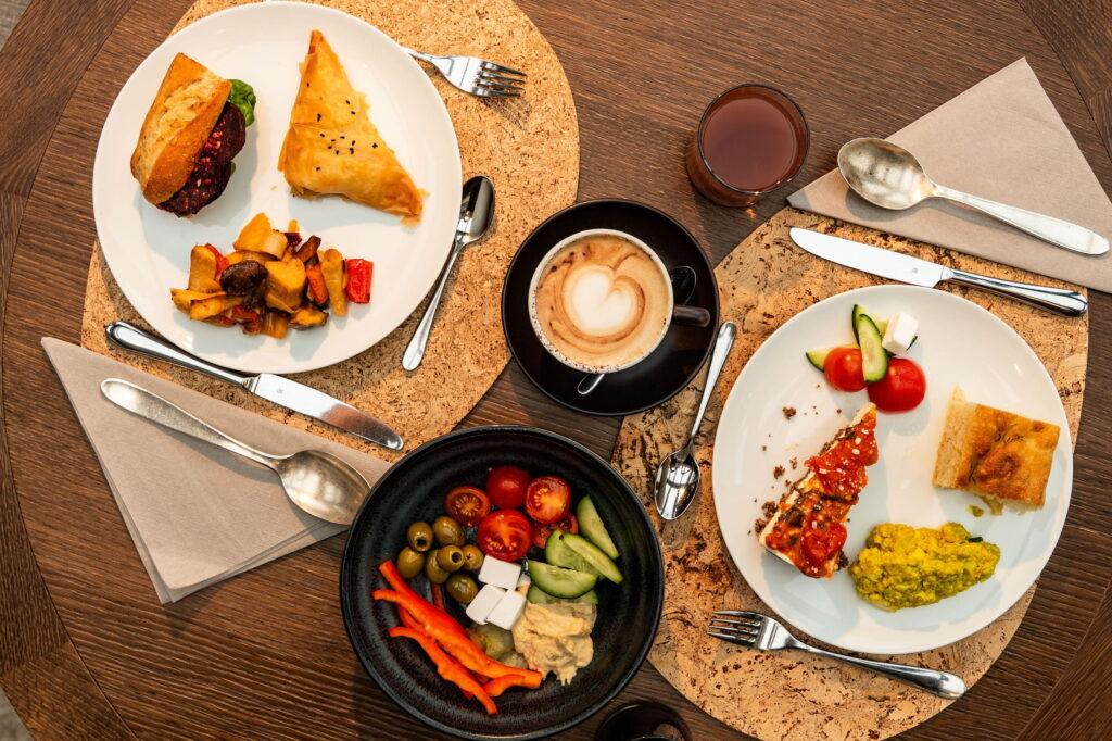Eine Draufsicht auf einen Holztisch, der mit verschiedenen veganen Speisen gedeckt ist, darunter ein Sandwich, Gebäck, gegrilltes Gemüse, ein Cappuccino und ein Salat mit Oliven, Hummus und Feta. Die Atmosphäre ist einladend und lebhaft im Paradiso Pure.Living Vegan Hotel in den Dolomiten.
