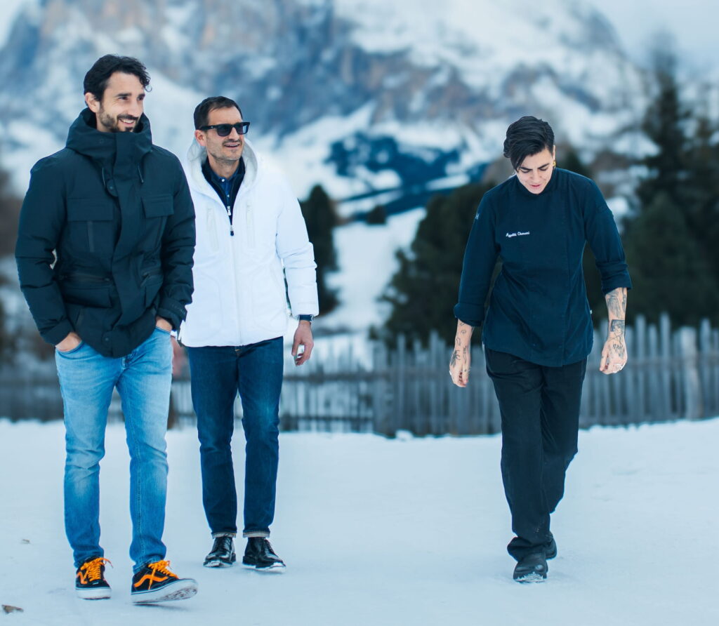 Die beiden Besitzer des veganen Hotels Paradiso Pure.Living spazieren zusammen mit Küchenchefin Aggeliki Charami in Winterkleidung durch eine verschneite Landschaft in Südtirol. Die beiden Männer links tragen dunkle und weiße Jacken, die Person rechts trägt eine dunkle Kochjacke. Im Hintergrund sind Berge und Bäume zu sehen, die eine ruhige, kühle Atmosphäre vermitteln.