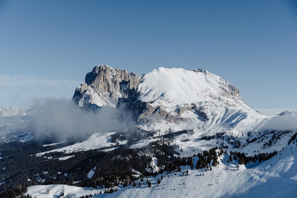 Schneebedeckter Berg der Dolomiten unter klarem blauem Himmel, mit vereinzelten Wolken und dichtem Wald darunter. Die Szene vermittelt Ruhe und natürliche Schönheit.