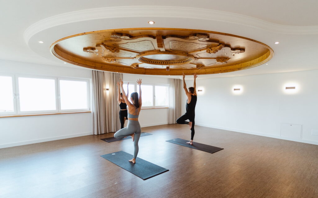 Drei Personen praktizieren Yoga in einem hellen, geräumigen Raum mit Holzboden im veganen Hotel Paradiso Pure.Living in den Dolomiten. Sie stehen auf Matten in der Baumhaltung unter einer kunstvollen Deckenverzierung und strahlen dabei ruhige Konzentration aus.