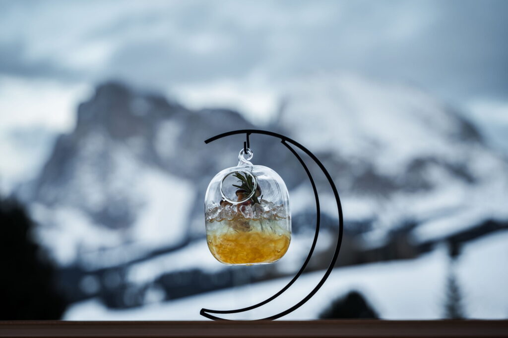Ein Getränk in einem terrarienartigen Glas mit Pflanzen und gelbem Boden auf einem halbmondförmigen Ständer, vor dem Hintergrund der schneebedeckten Berge der Dolomiten unter einem bewölkten Himmel.