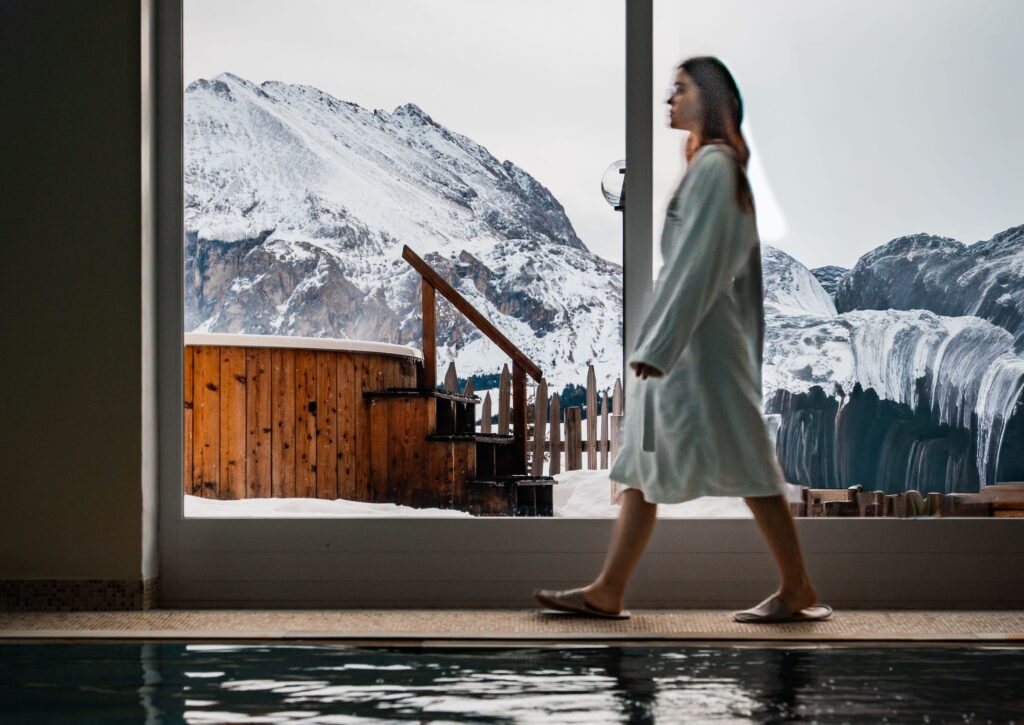 Eine Person in einem weißen Gewand geht in einem Innenraum in der Nähe eines Pools spazieren, während durch große Fenster schneebedeckte Berge der Dolomiten und ein hölzerner Whirlpool zu sehen sind, was eine ruhige, gemütliche Atmosphäre schafft.