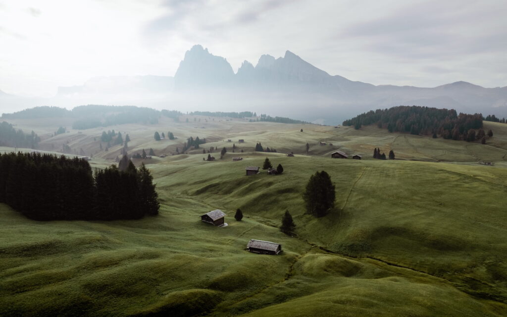Weite grüne Wiese auf der Seiser Alm in den Dolomiten in Südtirol mit verstreuten Holzhütten, umgeben von dunklen Bäumen. Im Hintergrund neblige Berge unter einem bewölkten Himmel, die Ruhe vermitteln.