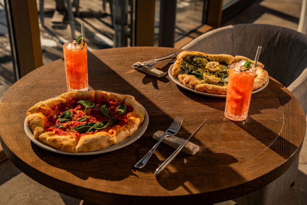 Zwei Pizzas stehen auf einem sonnenbeschienenen Holztisch im veganen Hotel Paradiso Pure.Living in Südtirol, eine mit roter Sauce und Basilikum, die andere mit Pesto. Daneben stehen zwei gekühlte Orangengetränke; das Besteck ist ordentlich gedeckt.