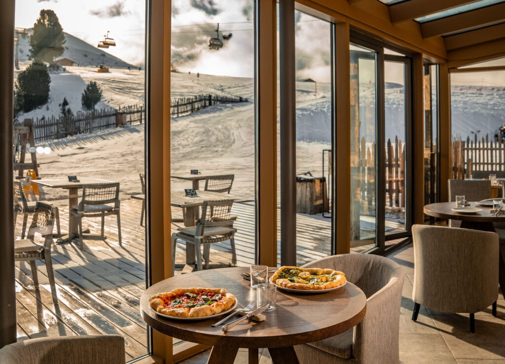 Gemütliches Restaurant des veganen Hotels Paradiso Pure.Living mit Blick auf schneebedeckte Berge durch Glaswände, mit zwei Pizzen auf einem Holztisch. Warme, einladende Atmosphäre, Skilift im Hintergrund.