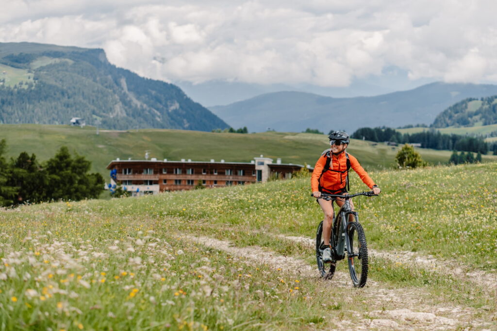 Radfahrer in orangefarbener Jacke fährt auf einem grasbewachsenen Weg in der malerischen der Dolomiten in Südtirol, mit dem entfernten veganen Hotel Paradiso Pure.Living im Hintergrund. Ruhige und abenteuerliche Stimmung.