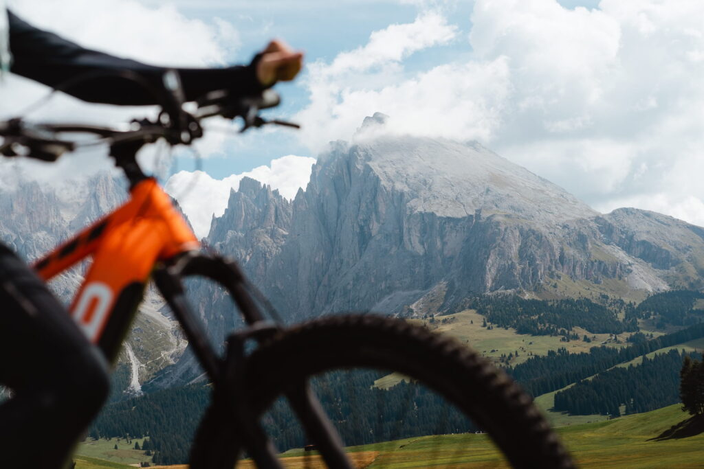 Eine Person auf einem Fahrrad konzentriert sich auf die majestätische Berglandschaft der Dolomiten im Hintergrund. Die Szene vermittelt Abenteuer und Ruhe unter einem teilweise bewölkten Himmel.