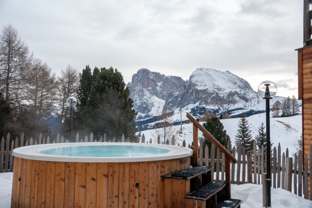 Holz-Whirlpool auf einer verschneiten Terrasse mit Stufen, umgeben von einem Lattenzaun. Im Hintergrund ragen Berggipfel der Dolomiten unter einem bewölkten Himmel empor und vermitteln Ruhe.