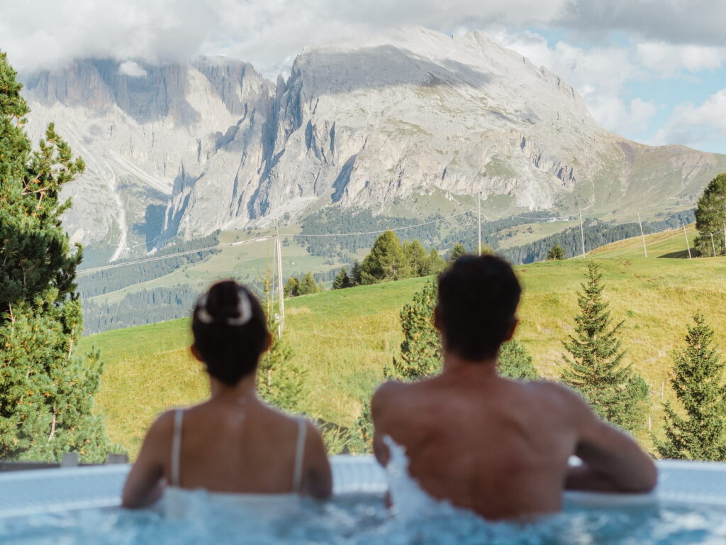 Ein Paar entspannt sich in einem Outdoor-Whirlpool im veganen Hotel paradiso Pure.Living und blickt auf die ruhige Berglandschaft der Dolomiten. Die üppig grünen Hügel und dramatischen Gipfel schaffen eine friedliche, idyllische Szenerie.