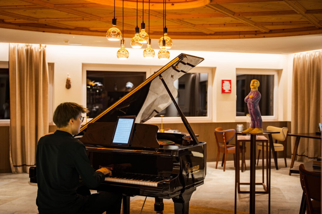 Eine Person in einem dunklen Hemd spielt auf einem Flügel in der gemütlichen Lobby des veganen Hotels Paradiso Pure.Living in den Dolomiten mit warmer Beleuchtung. Eine farbenfrohe Skulptur steht auf einem hohen Tisch in der Nähe und verleiht dem Raum eine künstlerische Note.