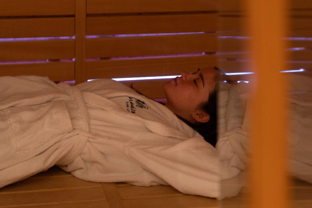Eine Person in einem weißen Bademantel entspannt sich in einer Holzsauna im Wellnessbereich des veganen Hotels Paradiso Pure.Living in Südtirol und liegt friedlich mit geschlossenen Augen da. Die warme, gemütliche Atmosphäre strahlt Ruhe und Behaglichkeit aus.