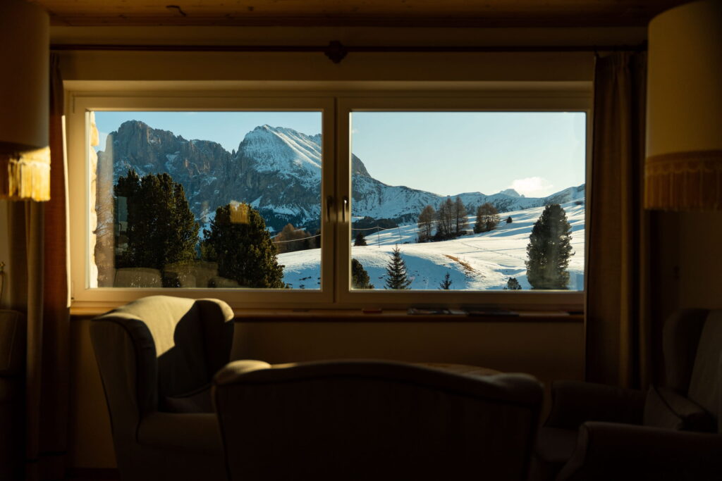 Blick durch ein Fenster auf schneebedeckte Berge und Bäume aus der gemütlichen Lobby im veganen Hotel Paradiso Pure.Living mit Sesseln. Die Szene vermittelt Wärme und Ruhe.
