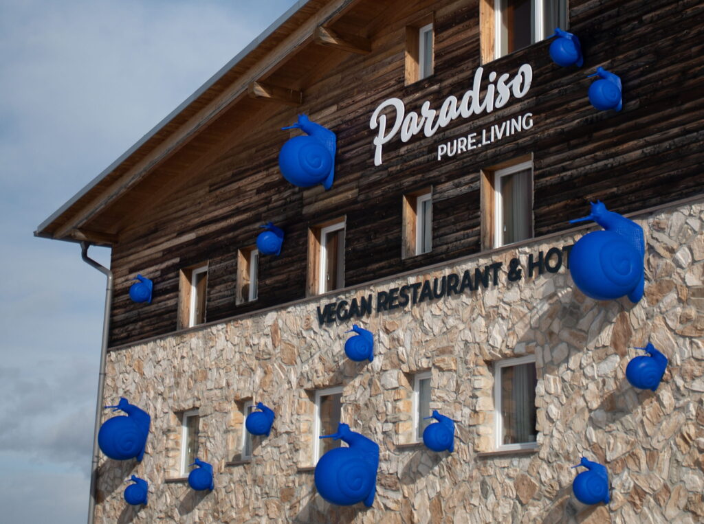 Alpines Gebäude mit den Schriftzügen „Paradiso Pure.Living” und „Vegan Restaurant & Hotel”. Große blaue Schneckenskulpturen zieren die Holz- und Steinfassade.