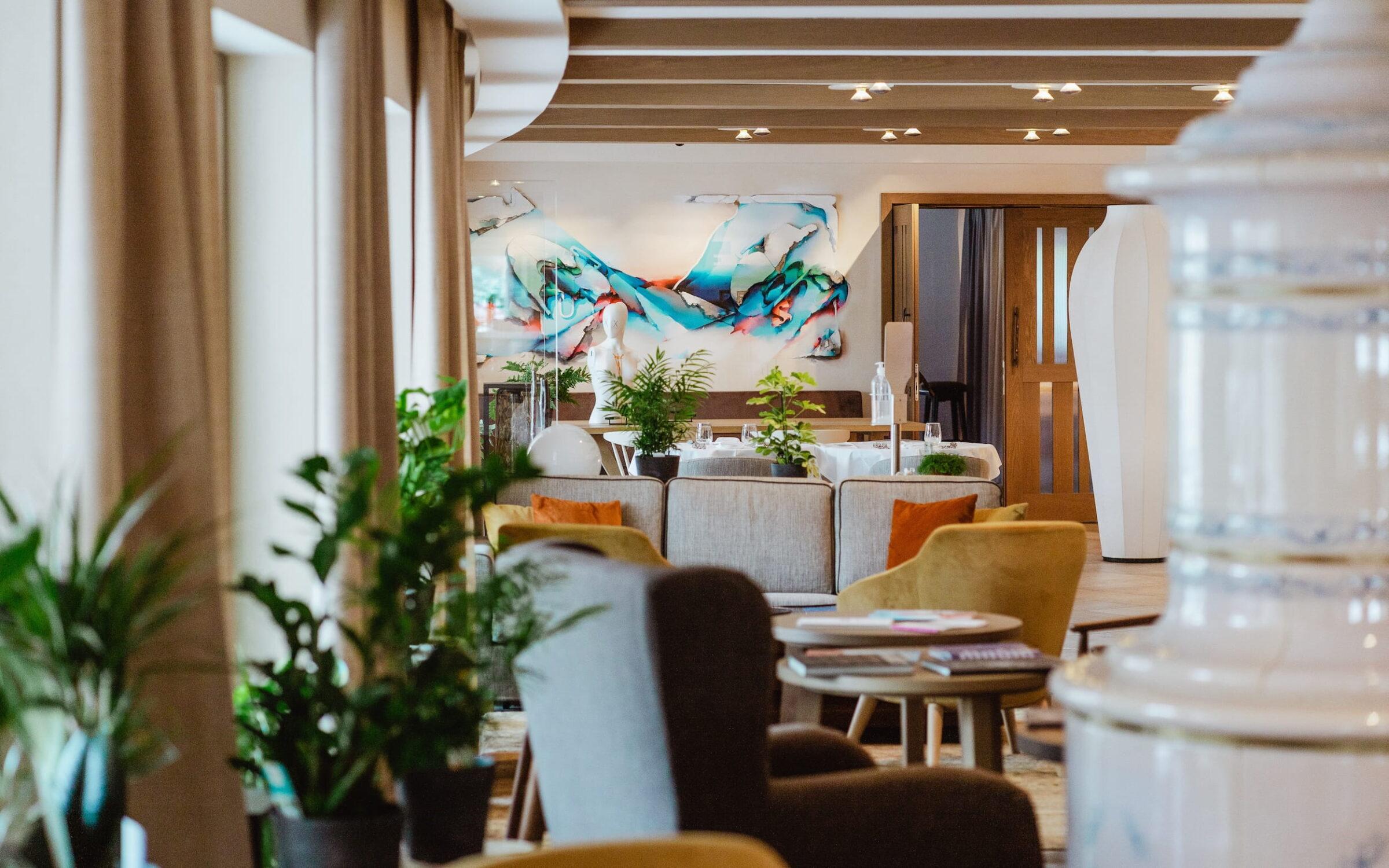Stilvolle Lounge im veganen Hotel Paradiso Pure.Living in den Dolomiten mit gemütlichen Sitzgelegenheiten, lebhaften abstrakten Kunstwerken an den Wänden und üppigen Grünpflanzen, die eine einladende, moderne Atmosphäre schaffen.