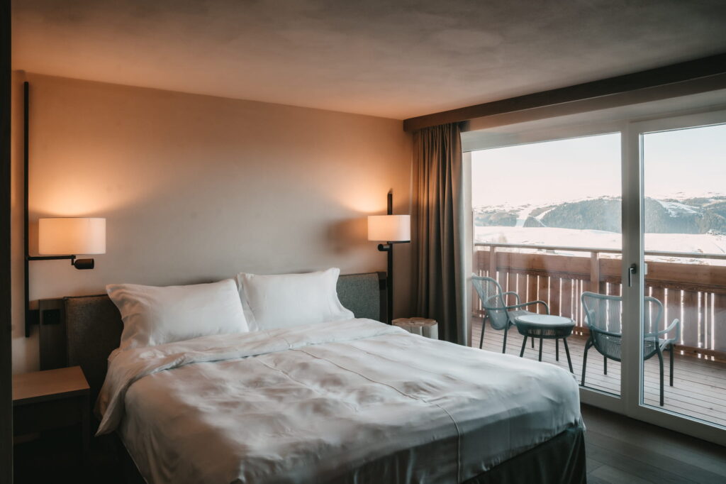 Gemütliches Hotelzimmer im veganen Hotel paradiso Pure.Living auf der Seiser Alm in Südtirol mit einem ordentlich gemachten Doppelbett, sanfter Beleuchtung durch Wandlampen, großen Fenstern mit Blick auf schneebedeckte Berge und einem Holzbalkon mit Stühlen.