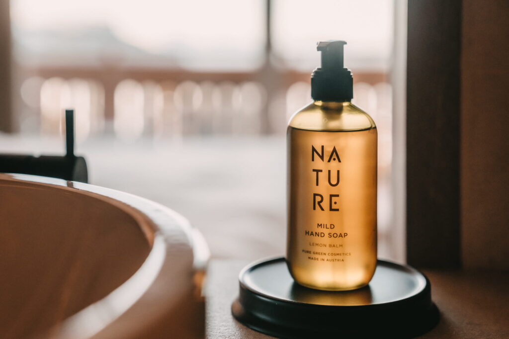 Bernsteinfarbene Flasche mit milder Handseife mit der Aufschrift „Nature“ auf einem runden Tablett neben einem Waschbecken aus Holz im veganen Hotel paradiso Pure.Living in Südtirol. Weichgezeichneter Hintergrund, warme und ruhige Atmosphäre.