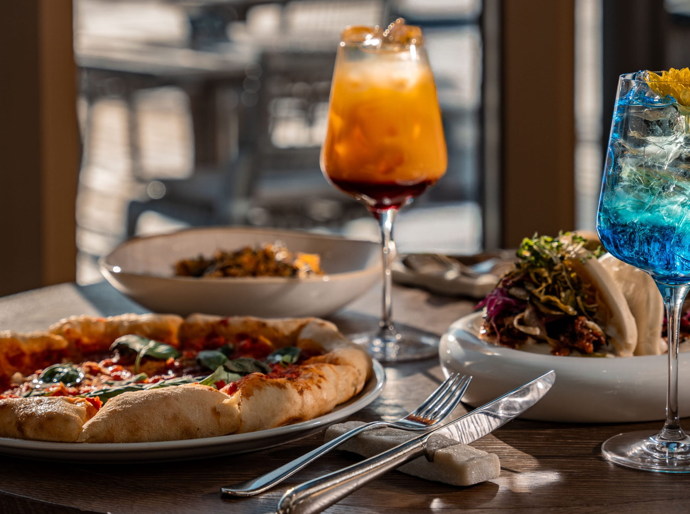 Eine bunte Auswahl an veganen Speisen und Getränken auf einem sonnendurchfluteten Tisch im veganen Hotel Paradiso Pure.Living in Südtirol, darunter frisch gebackene Pizza, Tacos, ein Orangen-Cocktail und ein blaues Getränk, sorgt für eine lebhafte Essensszene.