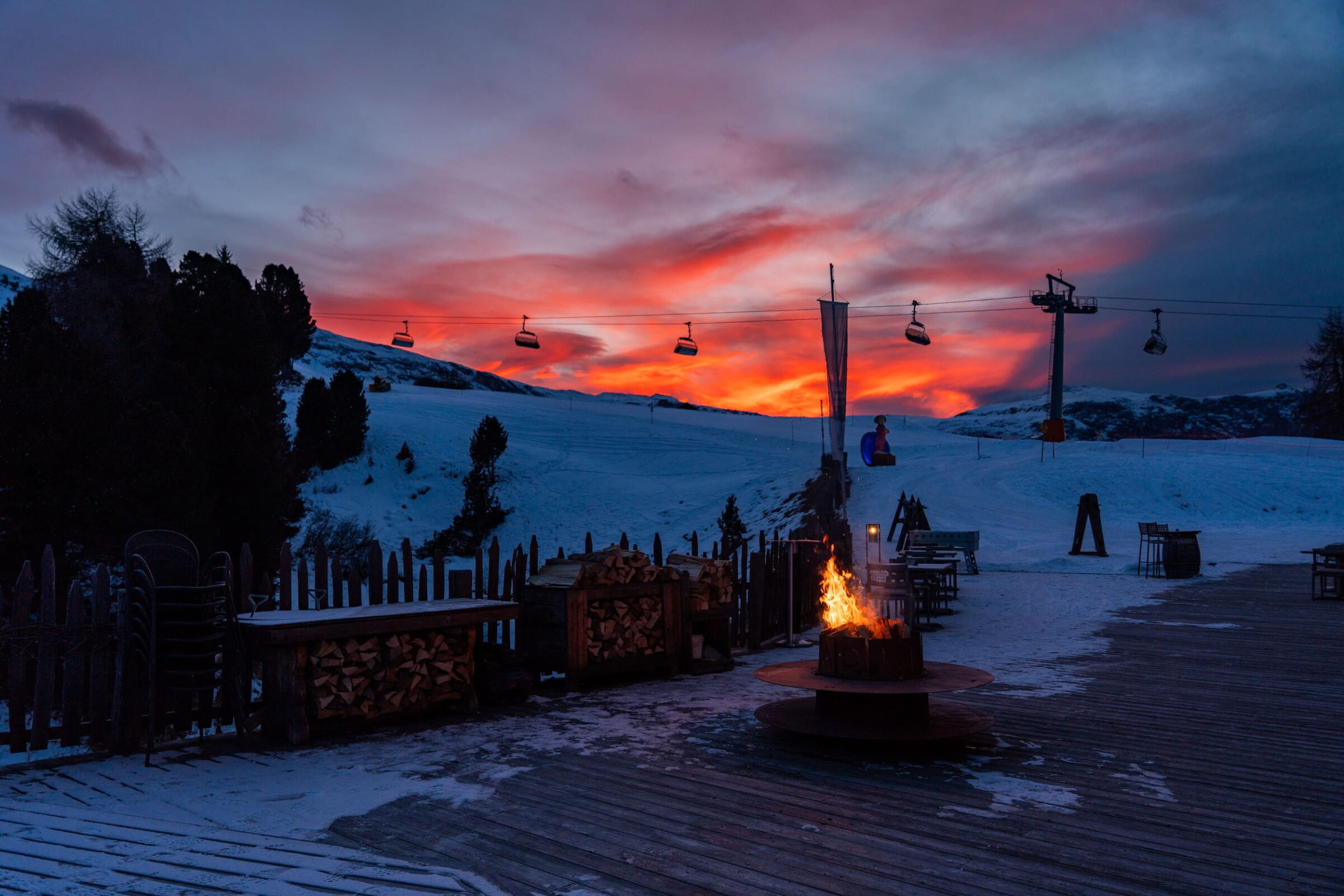 Eine verschneite Berglandschaft der Dolomiten in der Nähe des veganen Hotels Paradiso Pure.Living bei Sonnenuntergang mit leuchtend orangefarbenem und blauem Himmel. Skiliftstühle hängen vor dem farbenfrohen Hintergrund, und eine gemütliche Feuerstelle verleiht der Szene Wärme und vermittelt einen ruhigen Winterabend.