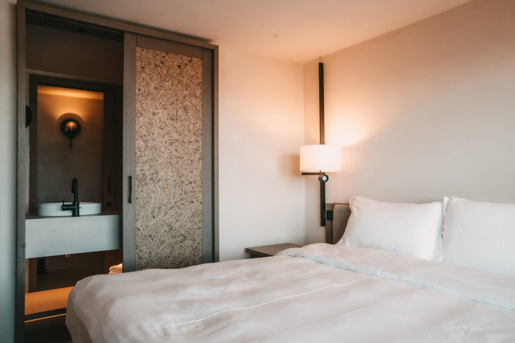 Ein gemütliches Hotelzimmer im veganen Hotel Paradiso Pure.Living verfügt über ein ordentlich gemachtes Bett mit weißer Bettwäsche. Die warme Beleuchtung einer Wandlampe und ein offenes Badezimmer mit einem Waschbecken aus Stein schaffen eine ruhige, moderne Atmosphäre.