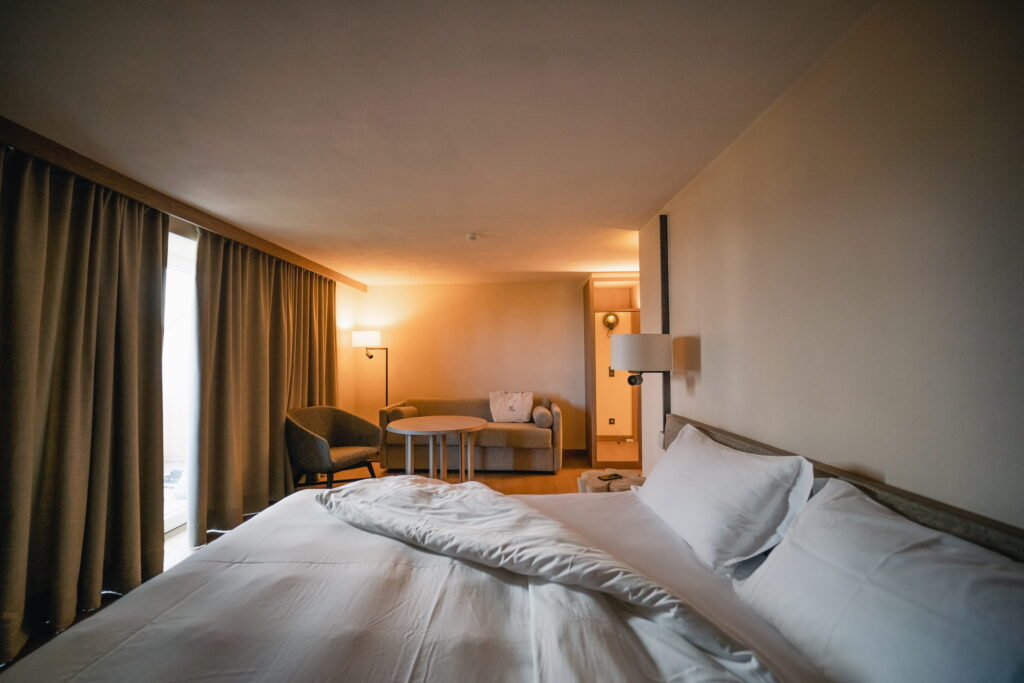 Modernes Hotelzimmer im veganen Hotel Paradiso Pure.Living in den Dolomiten mit einem ordentlich gemachten Doppelbett, sanfter Beleuchtung und einer gemütlichen Sitzecke. Dicke Vorhänge bedecken das Fenster und schaffen eine ruhige, einladende Atmosphäre.