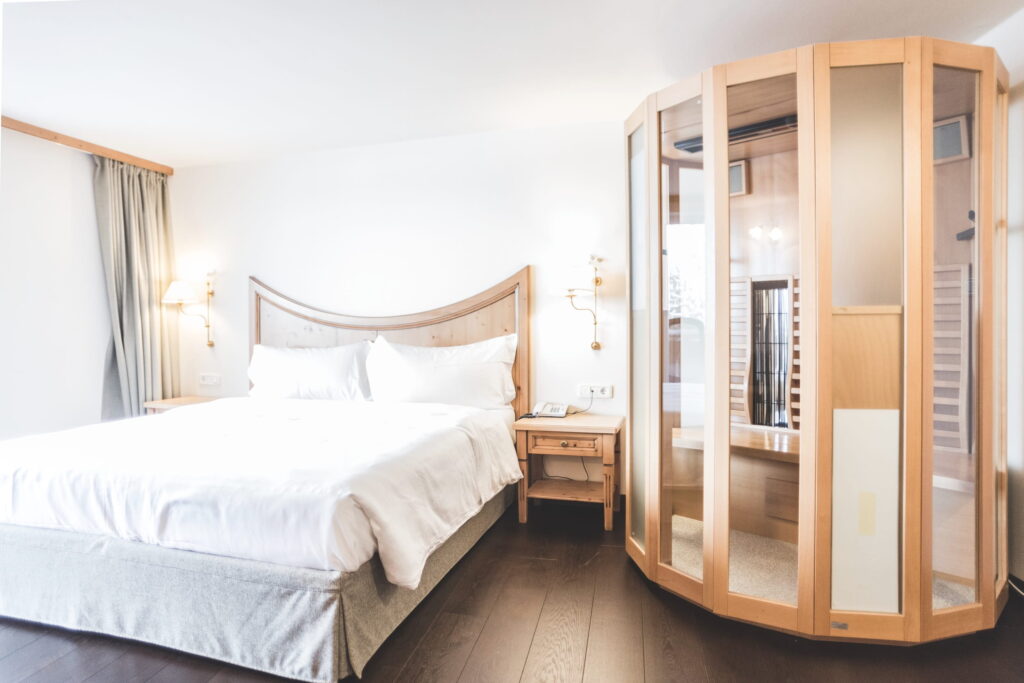 Ein helles, modernes Schlafzimmer im veganen Hotel Paradiso Pure.Living in Südtirol mit einem großen Bett mit weißer Bettwäsche, einem Kopfteil aus Holz und einem Beistelltisch. Neben dem Bett befindet sich eine Sauna aus Holz mit Glaswänden, die ein Spa-ähnliches Ambiente schafft.
