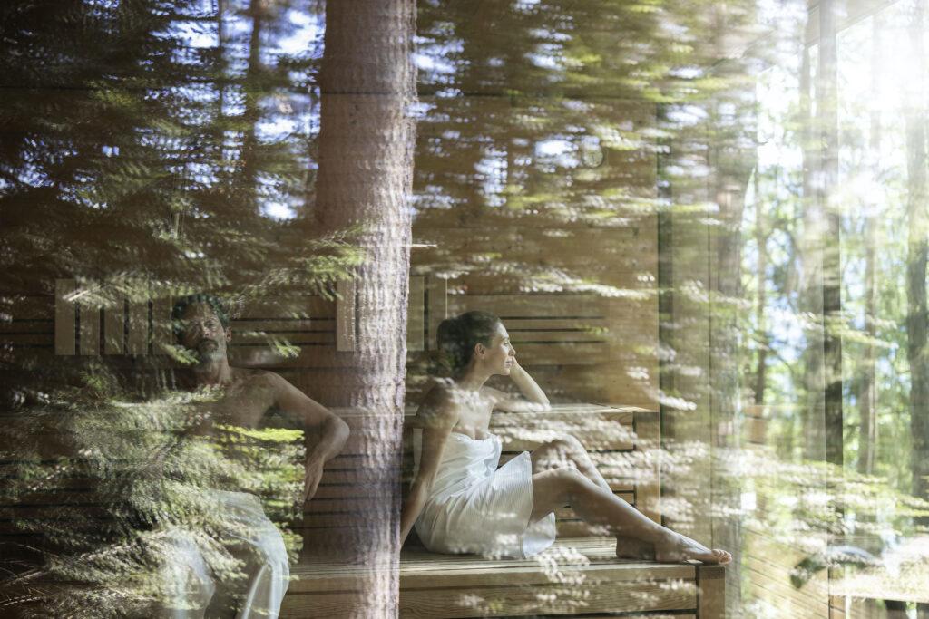 Junge Frau sitzt in der Waldsauna der nachhaltigen ADLER Lodge RITTEN. Im Fenster spiegeln sich die Bäume