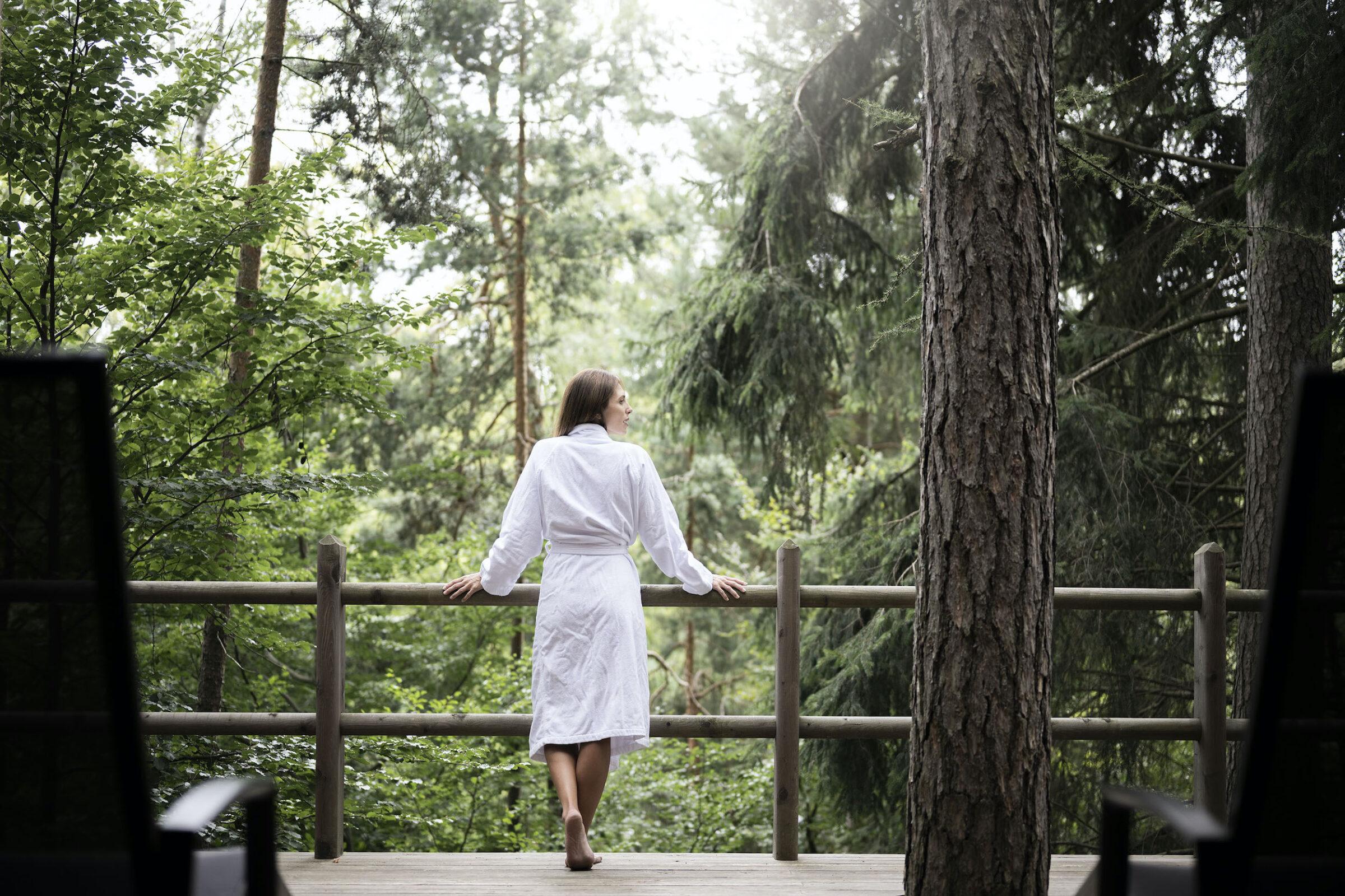 Nachhaltige ADLER Lodge RITTEN: Junge Frau steht im weißen Bademantel auf der Terrasse des Waldspas und schaut in die Bäume