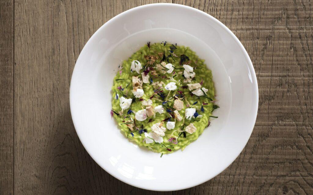 Eine weiße Schüssel auf einem Holztisch enthält grünes Risotto, garniert mit leuchtenden essbaren Blüten und zarten weißen Dekorationen. So werden die nachhaltigen kulinarischen Köstlichkeiten im ADLER Lodge RITTEN in Südtirol elegant und frisch präsentiert.