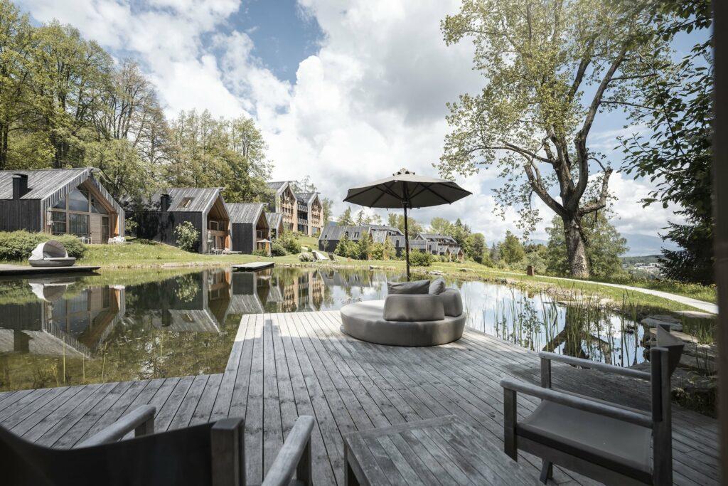 Malerischer Blick auf den See, in dem sich die Holzchalets der nachhaltigen ADLER Lodge RITTEN spiegeln. Liegestühle und ein Sonnenschirm laden zum Entspannen ein. Üppige Bäume unter einem teilweise bewölkten Himmel. Friedlich.