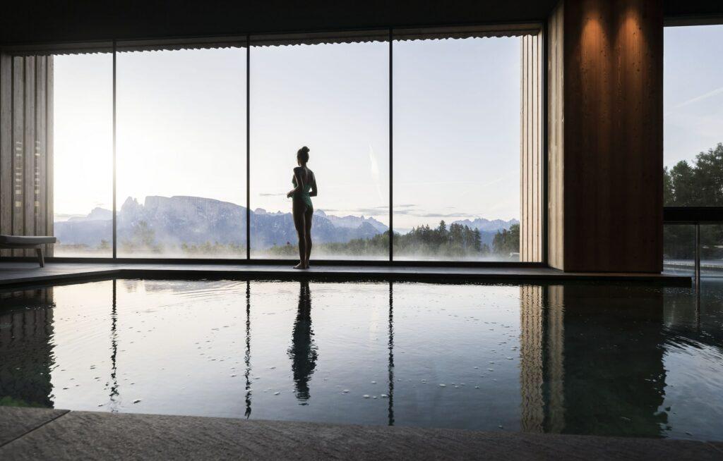 Eine Frau steht als Silhouette vor großen Glasfenstern mit Blick auf einen ruhigen Pool, während die Dolomiten im Hintergrund und der neblige Morgenhimmel eine friedliche Atmosphäre schaffen.