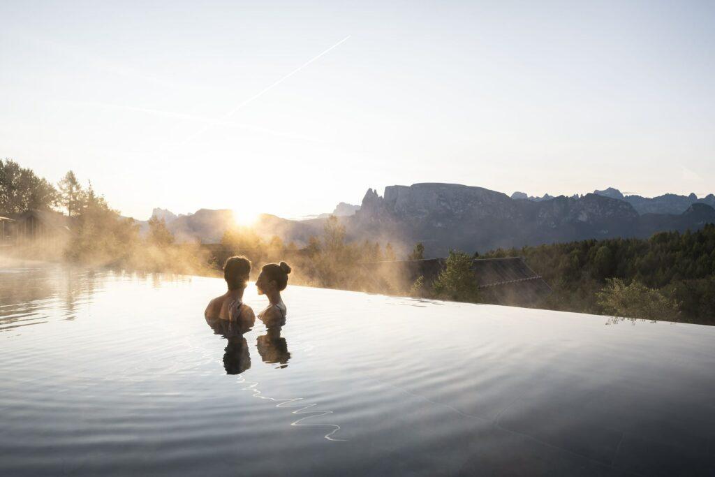 Ein Paar genießt einen ruhigen Moment im Infinity-Pool der nachhaltigen ADLER Lodge RITTEN bei Sonnenuntergang, während Nebel aufsteigt und malerische Berge und Bäume im Hintergrund zu sehen sind. Die Atmosphäre ist friedlich.
