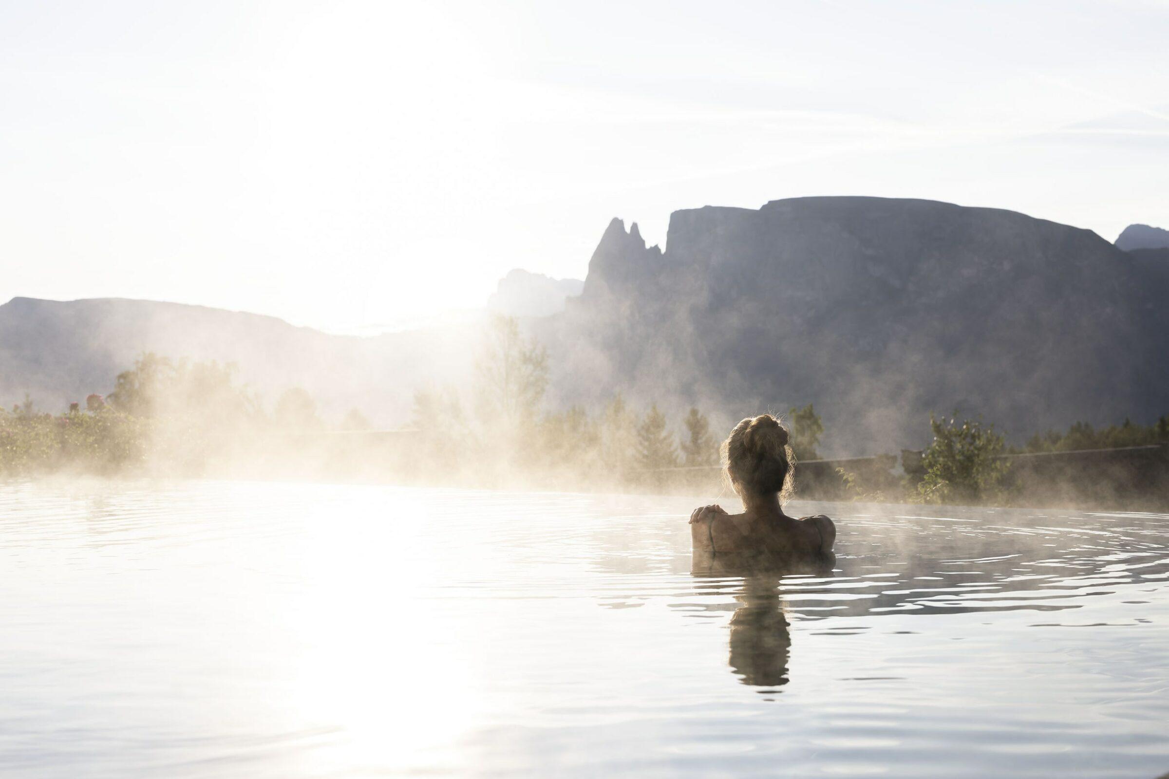 Eine Frau entspannt sich im dampfenden Infinity-Pool der nachhaltigen ADLER Lodge RITTEN vor einer bergigen Kulisse (Dolomiten). Die Morgensonne taucht alles in ein sanftes Licht und schafft eine ruhige, friedliche Atmosphäre.