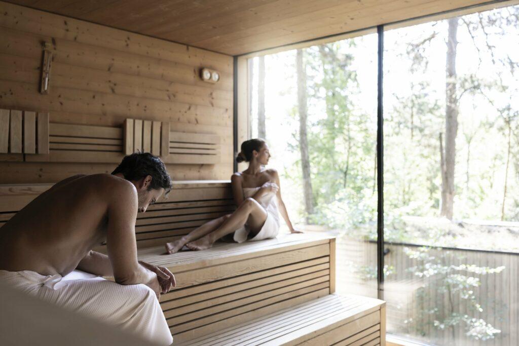 Zwei Menschen, in Handtücher gehüllt, sitzen in der ruhigen Holzsauna des ADLER SPA. Einer blickt durch ein großes Fenster auf hohe Bäume und schafft so eine ruhige, besinnliche Stimmung.