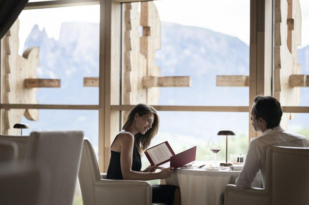 Ein Paar sitzt an einem Esstisch an einem großen Fenster mit Blick auf die Berge in der luxuriösen und nachhaltigen ADLER Lodge RITTEN. Die Frau lächelt auf eine Speisekarte; die beiden Gläser Rotwein lassen auf eine romantische Atmosphäre schließen.