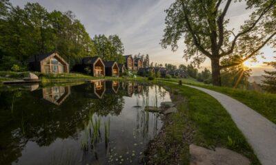Die Chalets der nachhaltigen ADLER Lodge RITTEN in Südtirol spiegeln sich bei Sonnenuntergang im ruhigen Wasser, umgeben von üppigen Bäumen. Ein Weg schlängelt sich entlang des Ufers und strahlt Ruhe und natürliche Schönheit aus.