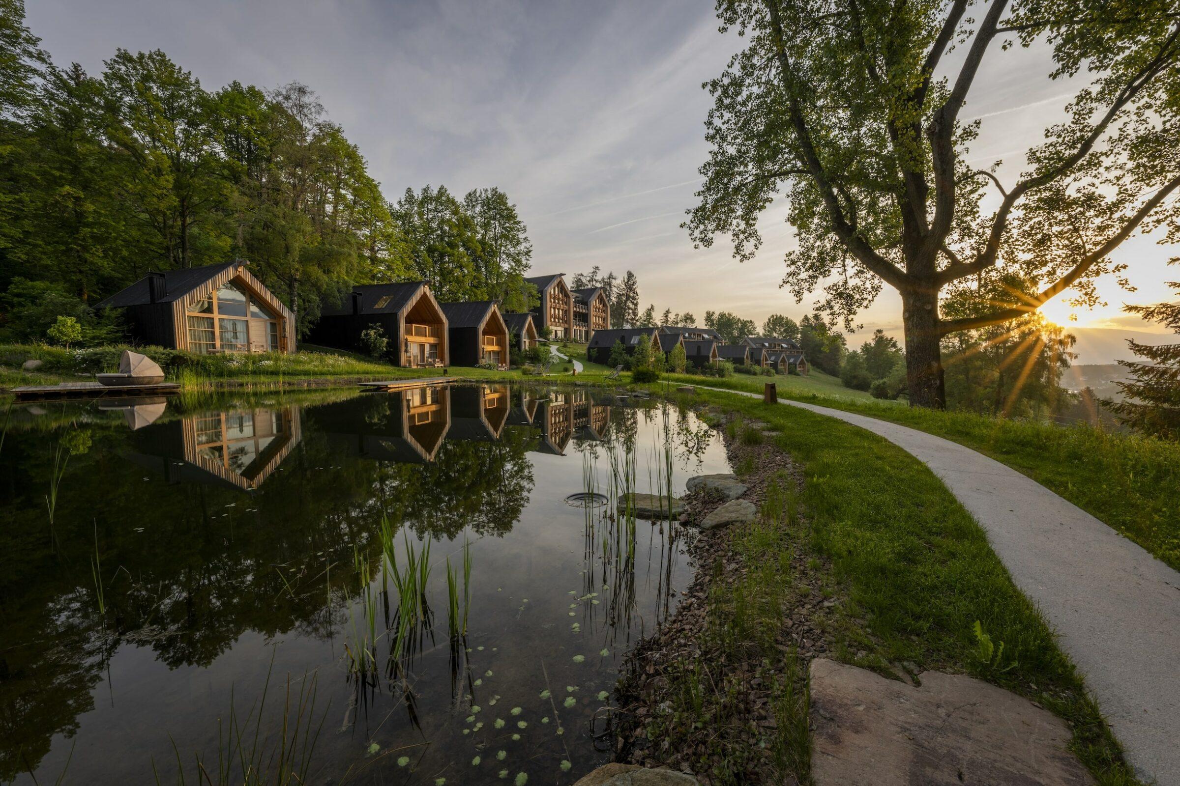 Die Chalets der nachhaltigen ADLER Lodge RITTEN in Südtirol spiegeln sich bei Sonnenuntergang im ruhigen Wasser, umgeben von üppigen Bäumen. Ein Weg schlängelt sich entlang des Ufers und strahlt Ruhe und natürliche Schönheit aus.