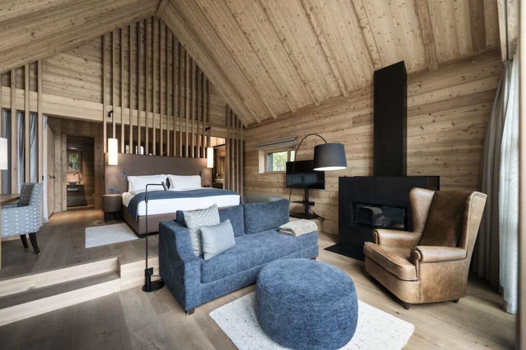Gemütliches Interieur mit Holzwänden und -decke in einem Junior-Chalet der nachhaltigen ADLER Lodge RITTEN. Mit großem Bett, Sitzecke mit blauem Sofa und Ottomane sowie Ledersessel am Kamin.