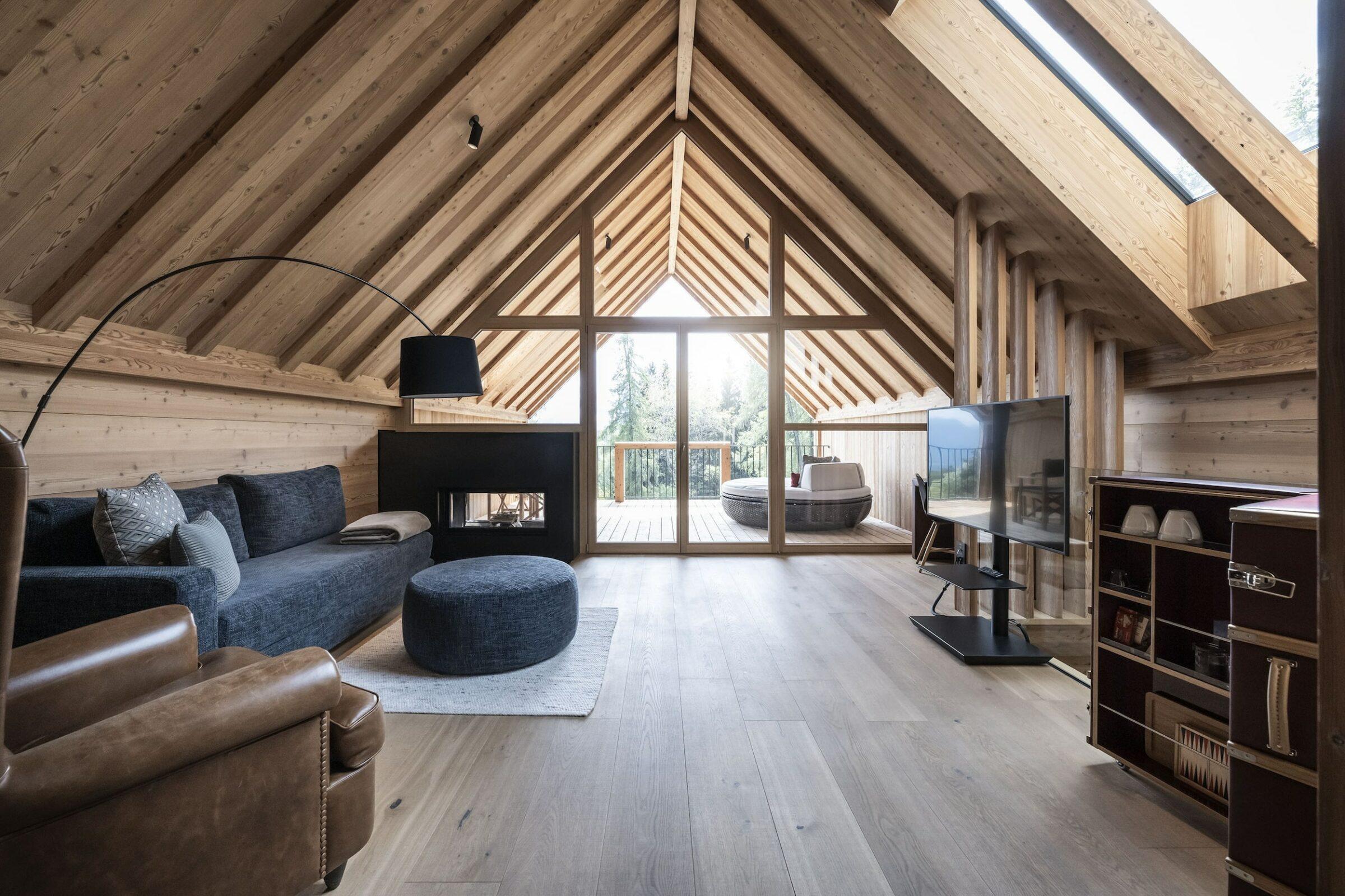 Geräumiges Wohnzimmer im Dachgeschoss mit Holzbalken und großen Fenstern in einem Chalet der nachhaltigen und luxuriösen ADLER Lodge RITTEN. Gemütliches graues Sofa, runder Ottoman, Ledersessel und ein Fernseher. Warme, einladende Atmosphäre.