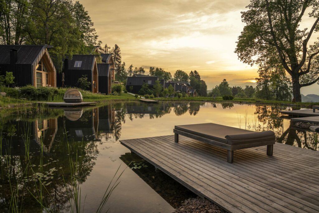 Eine ruhige Seestattung bei Sonnenuntergang, in der die modernen Holzchalets der ADLER Lodge RITTEN zwischen Bäumen liegen. Ein Steg mit einer Bank ragt über das ruhige Wasser, in dem sich der goldene Himmel über Südtirol spiegelt.
