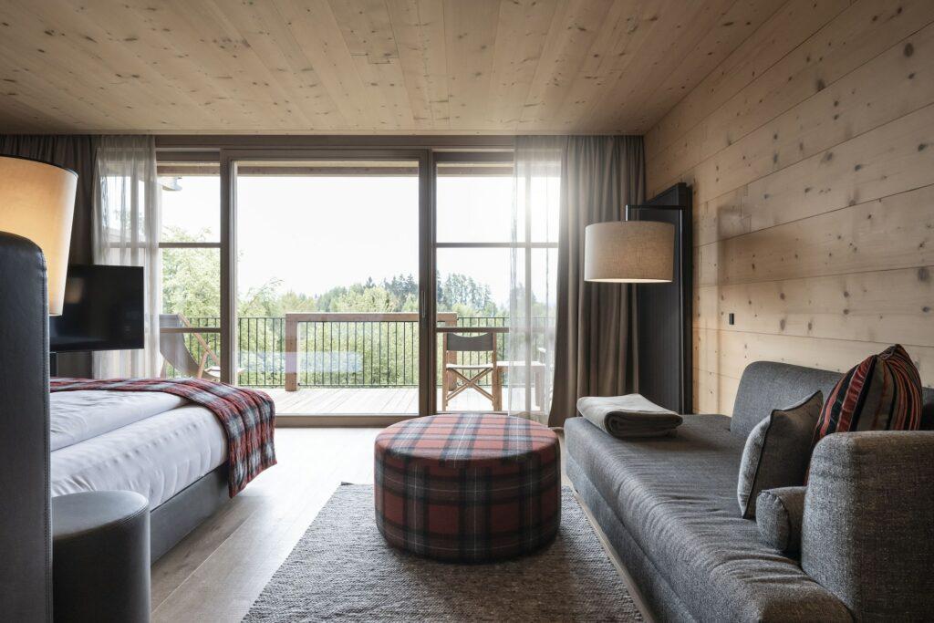 Eine gemütliche, moderne Junior Suite in der ADLER Lodge RITTEN in Südtirol verfügt über ein Bett und ein Sofa mit karierten Akzenten, Holzwände und ein großes Fenster mit ruhigem Blick auf den Wald. Warme, einladende Atmosphäre.