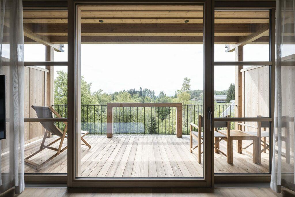 Durch die großen Glastüren der Junior Suite der ADLER Lodge RITTEN blickt man auf eine großzügige Holzterrasse mit Stuhl und Tisch. Der Blick auf die üppig grünen Bäume unter einem klaren Himmel vermittelt Ruhe und Entspannung.