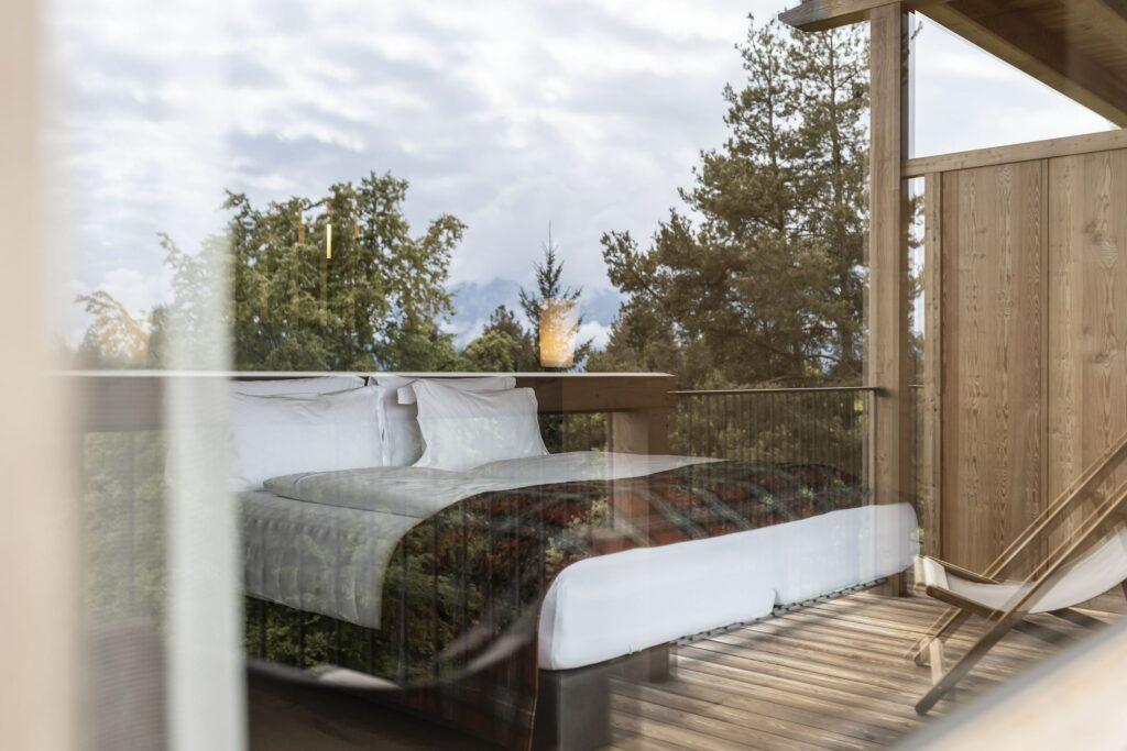 Das gemütliche Schlafzimmer der Junior Suite (ADLER Lodge RITTEN) mit einem großen Bett, das durch ein Glasfenster zu sehen ist und von Bäumen umgeben ist. Die Szenerie wirkt friedlich und lädt zum Entspannen inmitten der Natur ein.