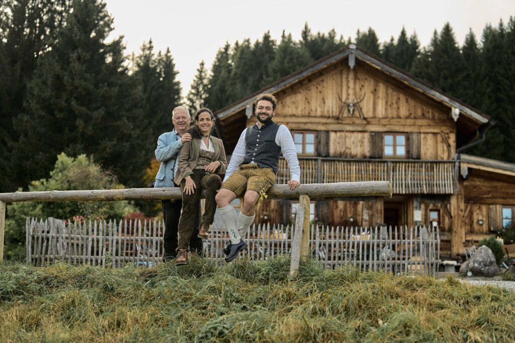 Die Inhaberfamilie Thurm des nachhaltigen Hotels Das Rübezahl in traditioneller bayerischer Tracht posiert fröhlich auf einem Holzzaun. Im Hintergrund sind ein rustikales Holzhaus und hohe Tannenbäume zu sehen.