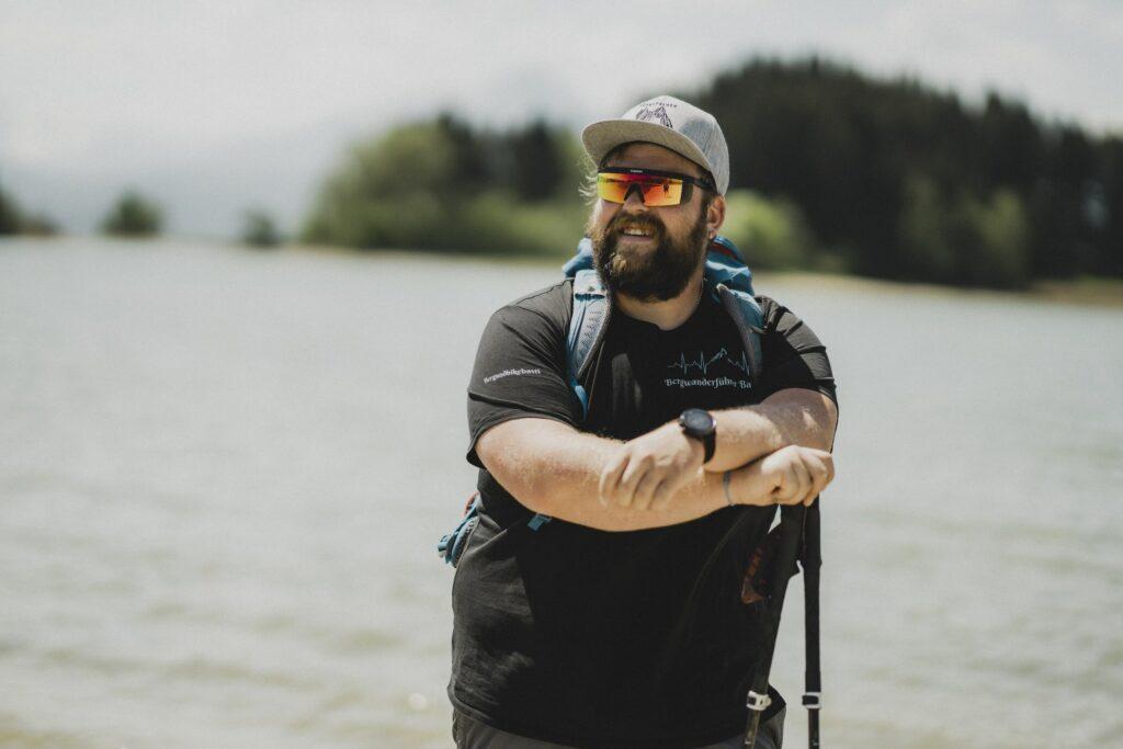Wander- und Berg-Guide des Hotel Das Rübezahl im Allgäu: Sebastian, ein Mann mit Bart und Sonnenbrille, steht an einem Bergsee, hält Trekkingstöcke in den Händen und trägt einen Rucksack und eine Mütze. Strahlender, sonniger Tag, Bäume im Hintergrund.